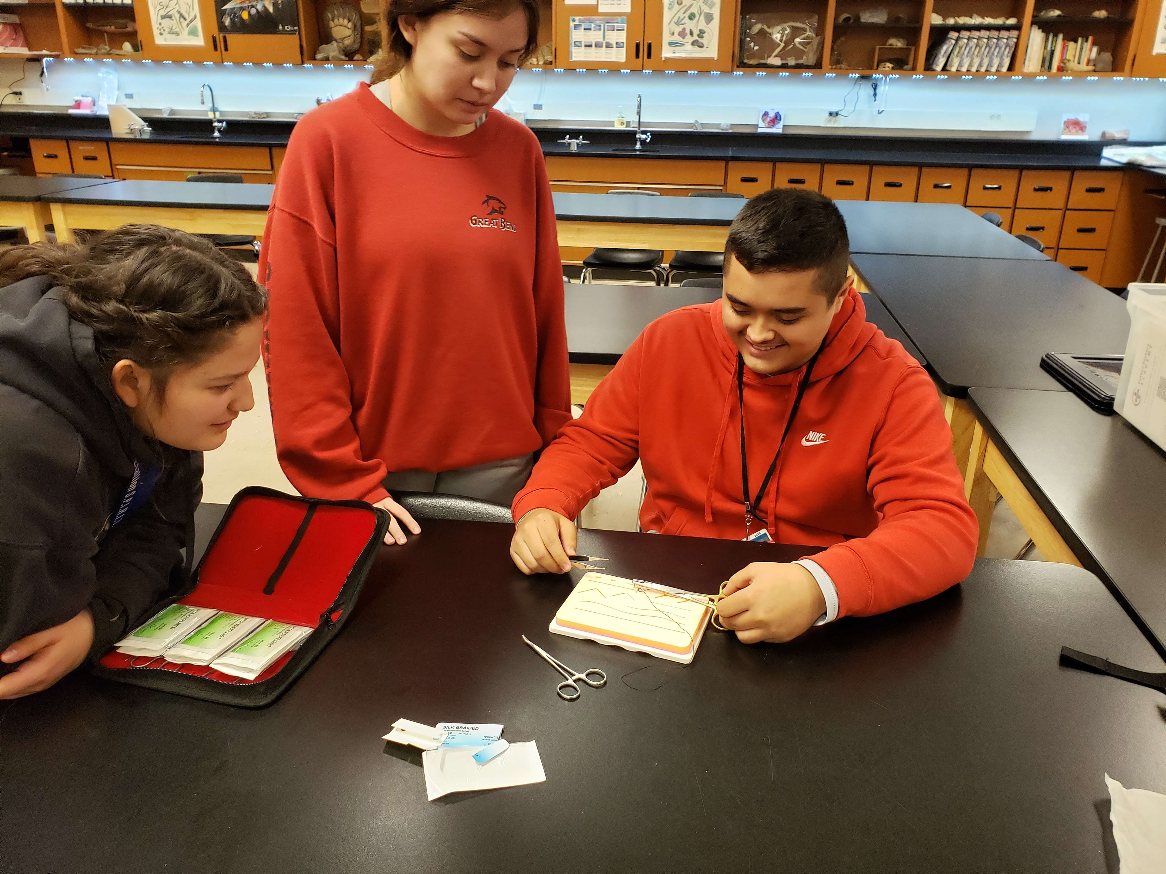 Great Bend High School students, Maria Valdovinos, Tresa Garcia, and Jason Scott, practice suturing techniques on a practice board. The board features different needle types and thread sizes to complete a variety of sutures. Another practice board for surgical knots is also available.