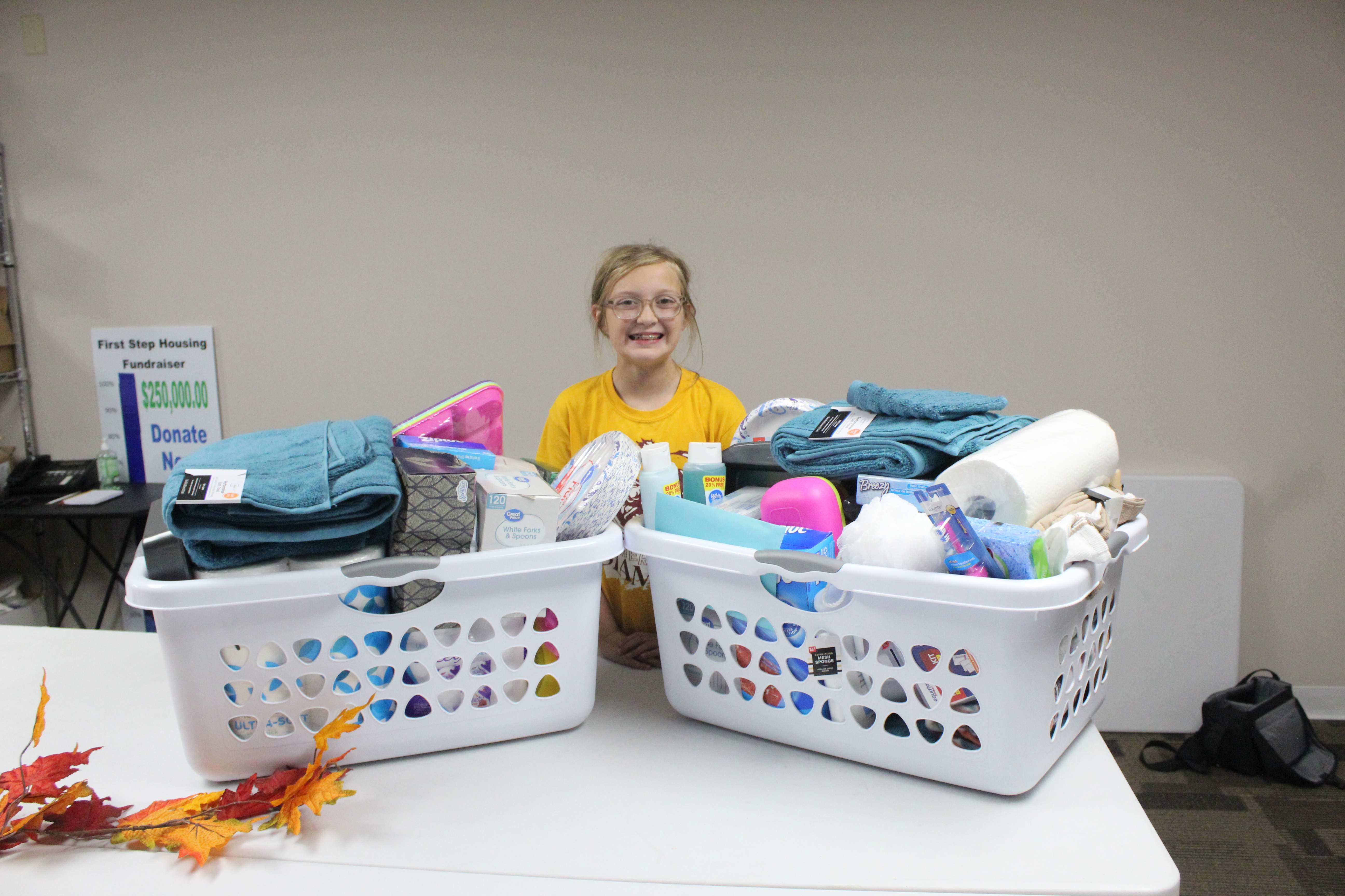 Kenzie Wolcott, representing Troop 11203, presents the First Step Housing program with two laundry baskets of household items for the project.<br>