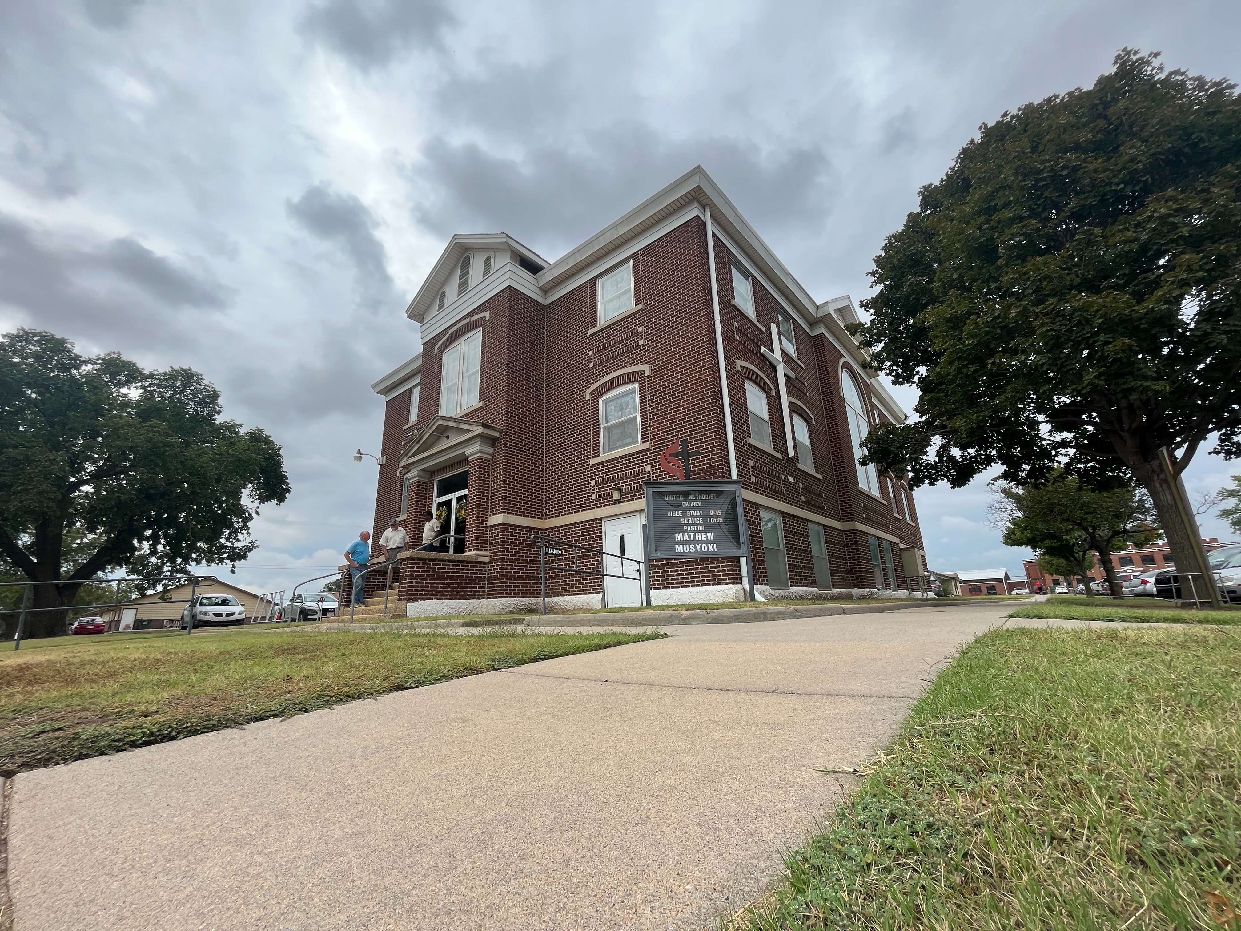 Cunningham Methodist Church in Cunningham, KS, with its pastor, Mathew Misyoki, next to its door talking to two congregates, Alan Albers (center) and Don Hellar (left). Photo by Olivia Bergmeier