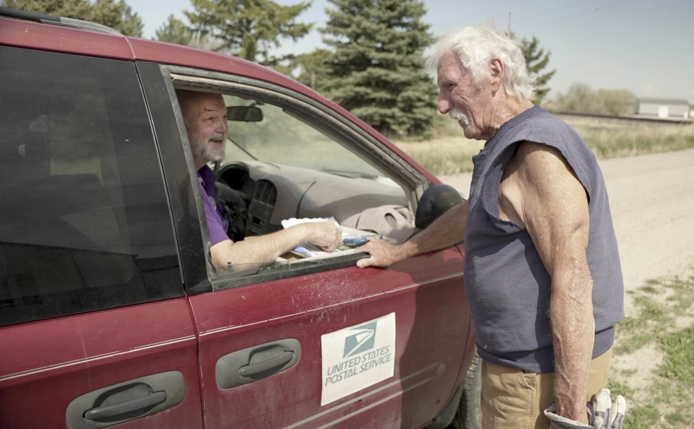 Rural letter carrier Roger McDonald, left, catches up with Edgar Clemens in rural Gering, Neb., on May 9, 2025 — McDonald’s first day back on the route after three months of medical leave. McDonald said he loves the sense of community fostered by the Postal Service, which he fears would disappear if his route was curtailed by cutbacks or eliminated by privatization. Photo by Nick Loomis/The Midwest Newsroom