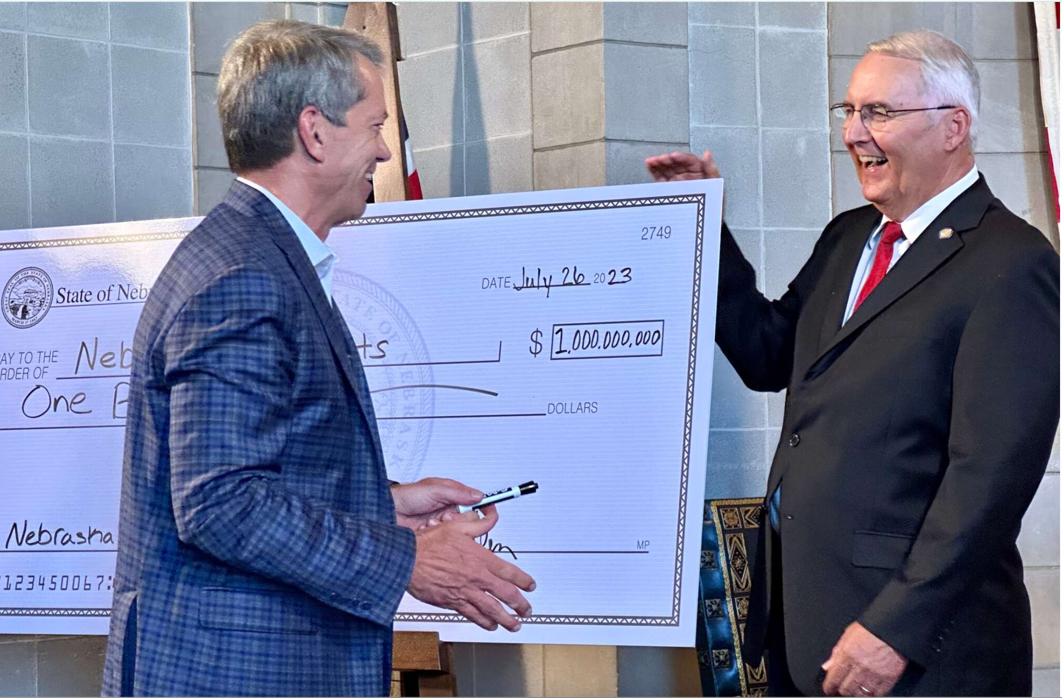 Nebraska Gov. Jim Pillen and State Sen. Rob Clements of Elmwood laugh after he signs a mock check signifying state investments in K-12 education. (Aaron Sanderford/Nebraska Examiner)
