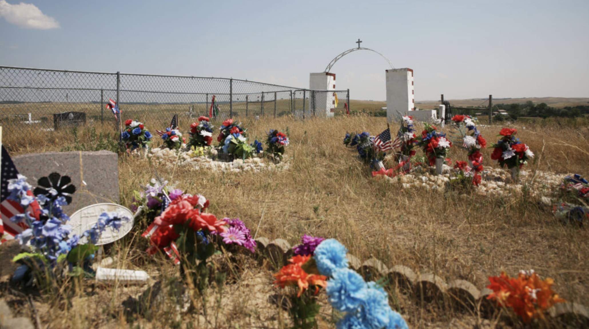 Plastic flowers and American flags are placed atop graves at the Wounded Knee Memorial and cemetery on June 30, 2024. (Makenzie Huber/South Dakota Searchlight)