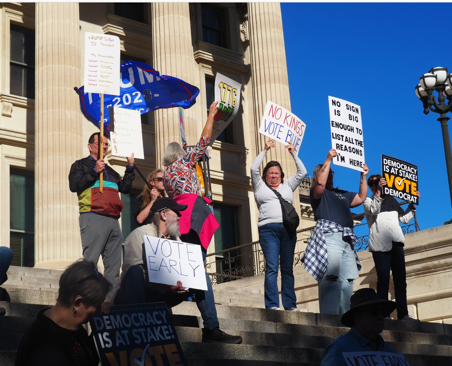 A contingent attending a rally Oct. 17, 2026, outside the Kansas Capitol in advance of the Nov. 3 election do their best to obscure the lone person waving a flag in support of President Donald Trump. The objective of the event organized by groups aligned with the No Kings movement is to encourage Kansans to vote in-person with an advance ballot. (Photo by Tim Carpenter/Kansas Reflector)