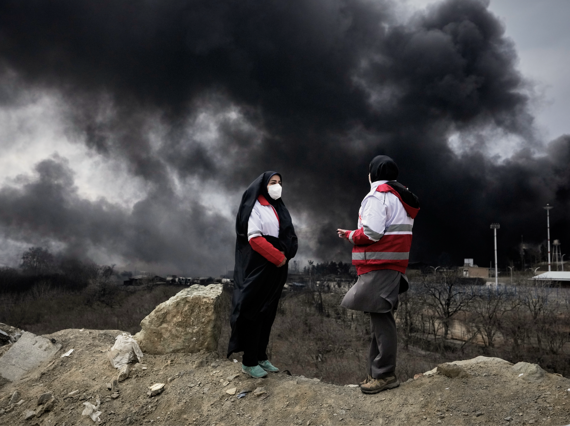Two women from the Iranian Red Crescent Society stand as a thick plume of smoke from a U.S.-Israeli strike on an oil storage facility late Saturday rises in the sky in Tehran, Iran, Sunday, March 8, 2026. (AP Photo/Vahid Salemi)