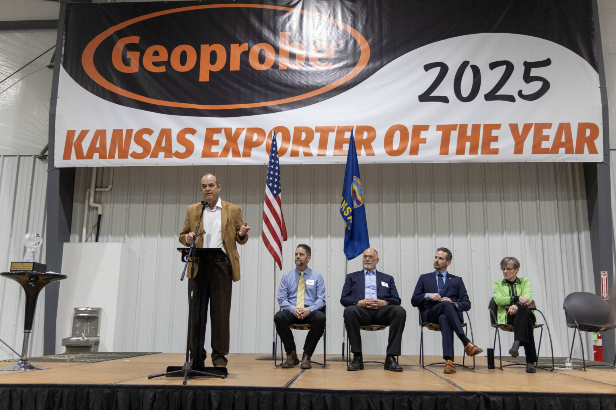 Omli speaks to crowd during "Exporter of the Year" award event, with the prize award to his right. <b>From left to right sitting down:</b> Tony Bowell, lead singer of Sunset Sinners,  Jeff Willis Chair of the Kansas International Trade Coordinating Council (KITCC), Joshua Jefferson, Deputy Secretary of Business Development for the Kansas Department of Commerce, (KDOC) Governor Kelly - Photo, Maria Gutierrez of Geoprobe Systems&nbsp;