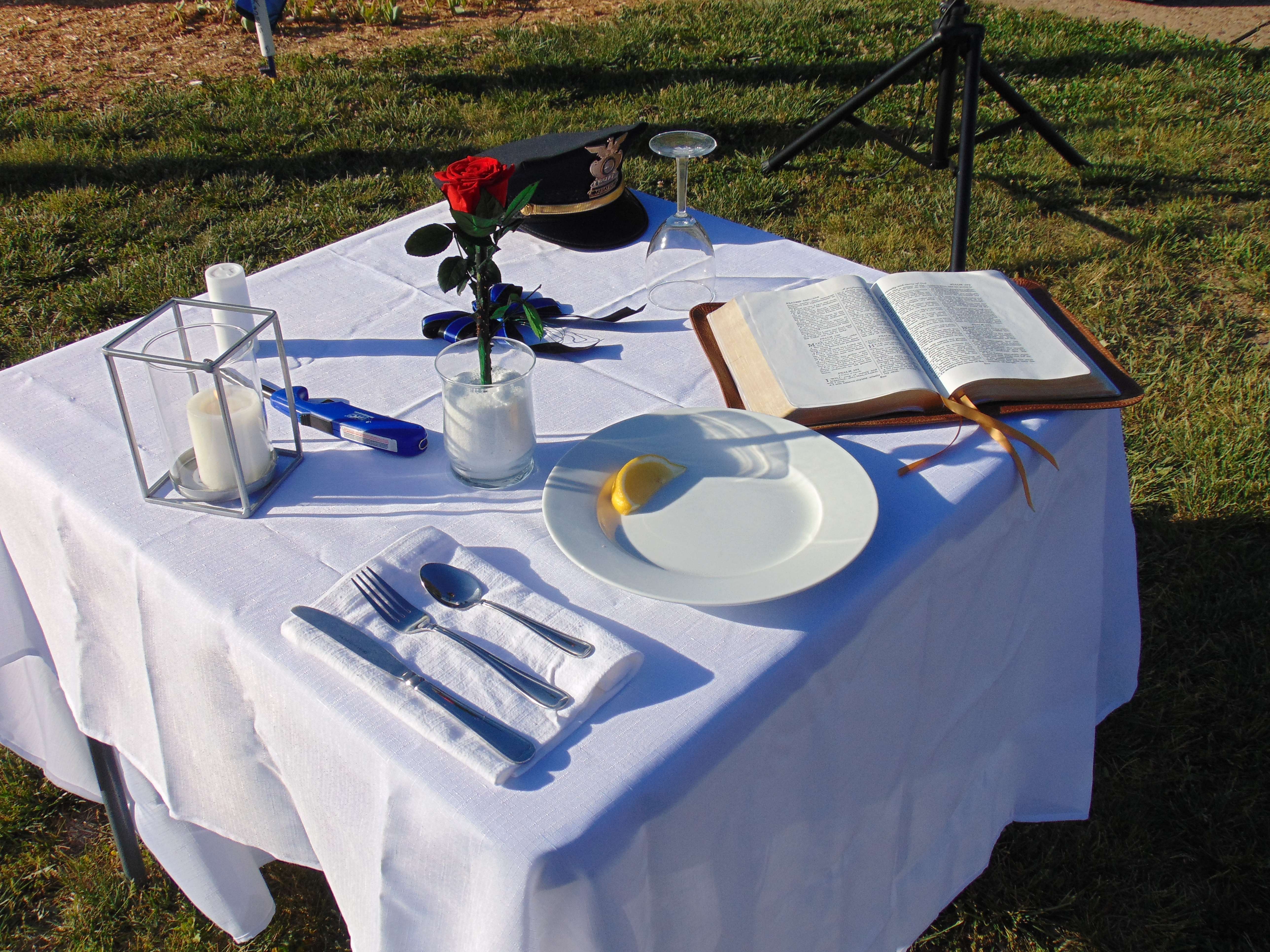 Meaning: <b>Square table;</b> Showing the many facets of first responders: father or mother, brother or sister, son or daughter and citizen hero. <b>White tablecloth;</b>&nbsp;symbolizes the purity of the motives when answering the alarm. <b>Rose; </b>represents the love for the fallen comrades and the sacrifice that was made to protect the innocent. <b>Bible open to Psalm 91; </b>represents the strength gained through faith in God who sustains and protects us. <b>Slice of lemon</b>; Reminds of the bitter taste after losing a comrade or a loved one. <b>Pinch of Salt</b>; represents the tears shed for fallen comrades. <b>Blue ribbon;</b>&nbsp;represents the trust in and loyalty to comrades present today. <b>Lit candle; </b>light of hope that all will return home safely tonight. <b>Inverted glass; </b>representing the fallen's inability to share a toast - Photo, Nicolas Fierro