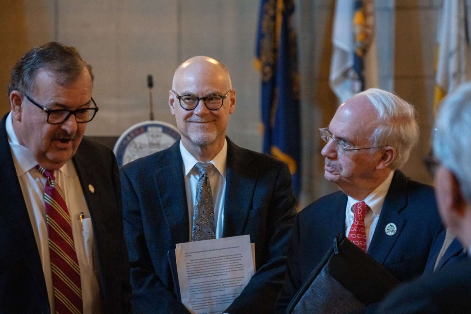 University of Nebraska President Dr. Jeffrey Gold, right, speaks with state senators and NU regents after a forum with dozens of Nebraska state senators regarding NU’s proposal to buy out the share of Nebraska Medicine owned by Clarkson Regional Health Services. At center is Dr. Bill Lydiatt, Clarkson’s CEO, and at left is State Sen. Mike Jacobson of North Platte. Jan. 14, 2026. (Zach Wendling/Nebraska Examiner)