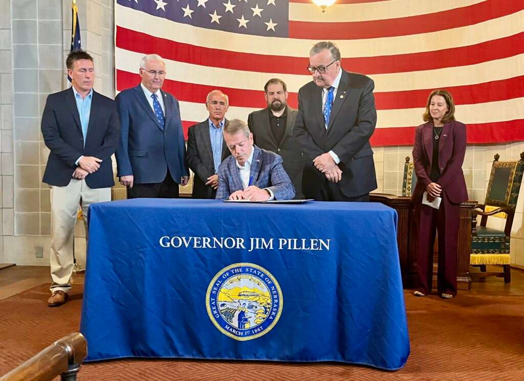  Gov. Jim Pillen signs executive order regulating data centers next to State Sen. Mike Jacobson of North Platte. Behind him are Director of Nebraska Department of Water, Energy and Environment Jesse Bradley, Sen. Rob Clements of Elmwood, Sen. Tony Sorrentino of the Elkhorn area, Sen. Eliot Bostar of Lincoln, and Director of the Nebraska Department of Economic Development Maureen Larsen. (Erin Bamer/ Nebraska Examiner)