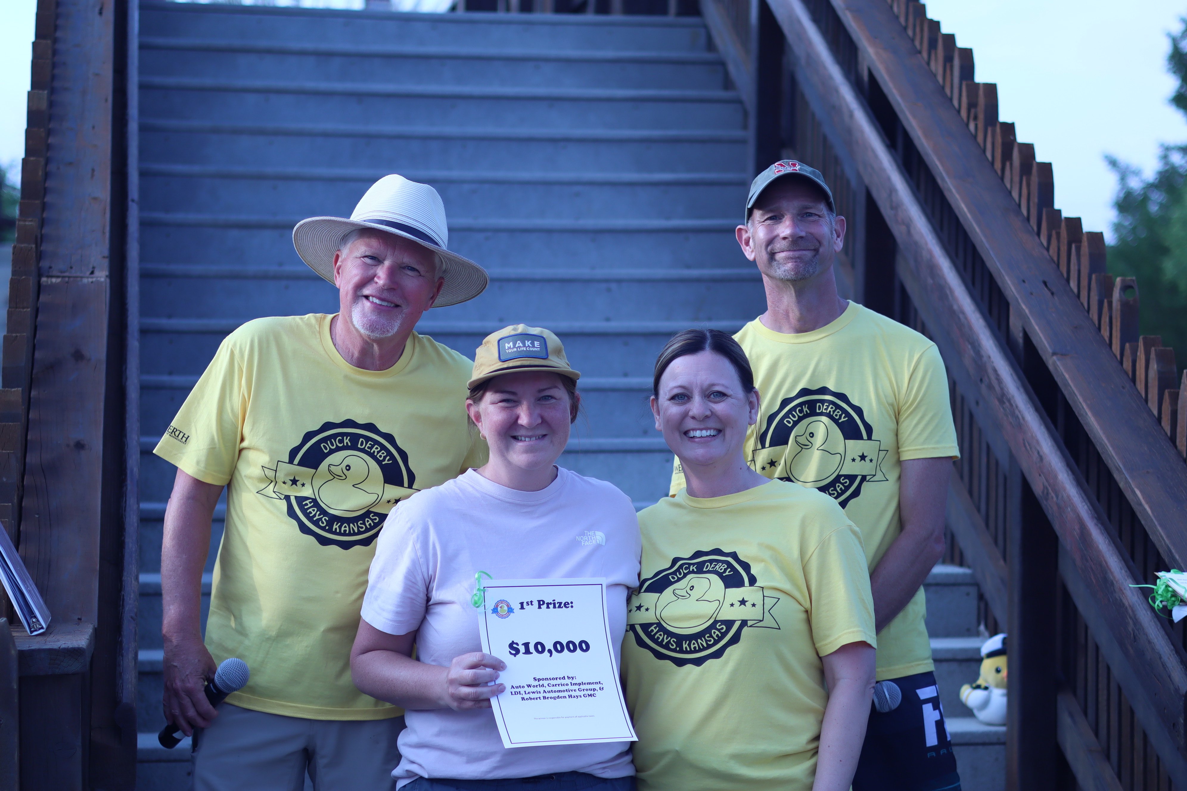 Brett Denham holding her $10,000 award for placing first at the 2026 Hays Duck Derby, standing next to MC Gary Shorman (left), Area Director for Kansas Big Brothers Big Sisters Jenny Bates (middle), and MC Mike Holliday (right). 