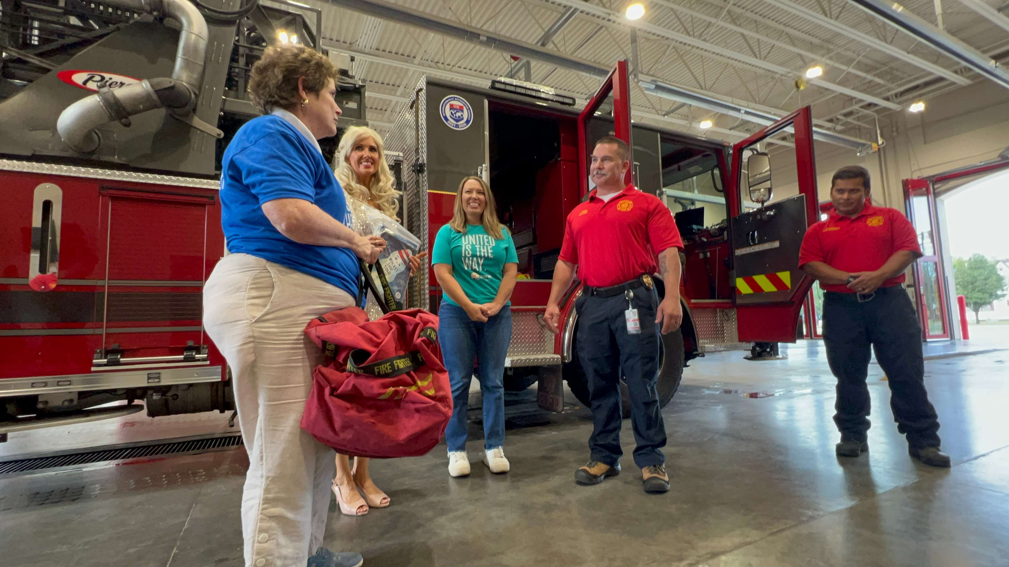 Elizabeth Walton, development manager for United Way of Reno County, hands a bag of books from the Dolly Parton Imagination Library to Hutchinson Fire Chief Jeremy Unruh on Friday, Sept. 25, 2026, at Fire Station No. 1. United Way of Reno County co-chair Christina Weber, dressed as Dolly Parton, President and CEO Lacey Mills, and Hutchinson Fire Department Community Risk Specialist and External Standards Manager Wade Mirasol look on. (Hutch Post Photo/Sean Boston)